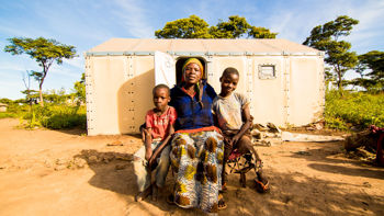 En mamma och två barn sitter framför en nödbostad och tittar in i kameran.
