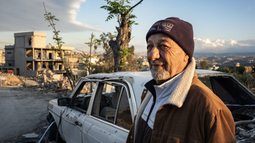 Mohammed, 80 år, står bredvid en krigsskadad bil i Tyros i södra Libanon. Mohammed flydde för sitt liv i september 2024, när en israelisk missil slog ner bara några meter från hans hem.