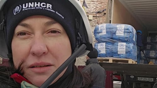 En kvinna med UNHCR-mössa står i vinterkläder framför en hjälpkonvoj i Ukraina.