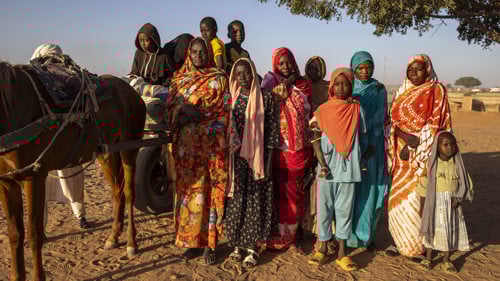 En grupp människor som flytt står i rad, de tittar allvarsamt in i kameran.