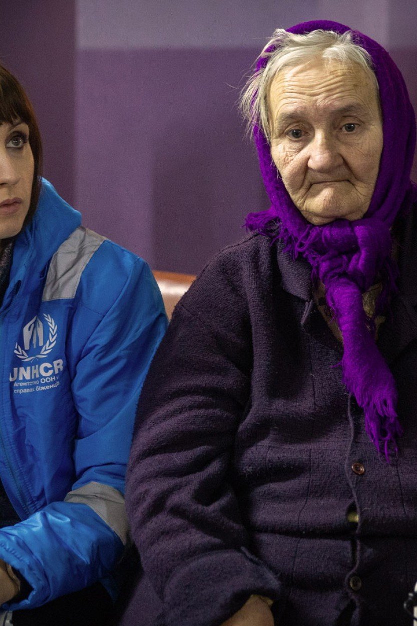 Liudmila, en äldre ukrainsk kvinna, sitter på en säng. Bredvid henne sitter en UNHCR-medarbetare i blå väst.