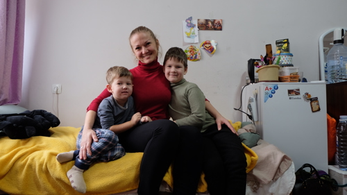 En kvinna sitter på en säng tillsammans med sina två barn. De ler mot kameran.