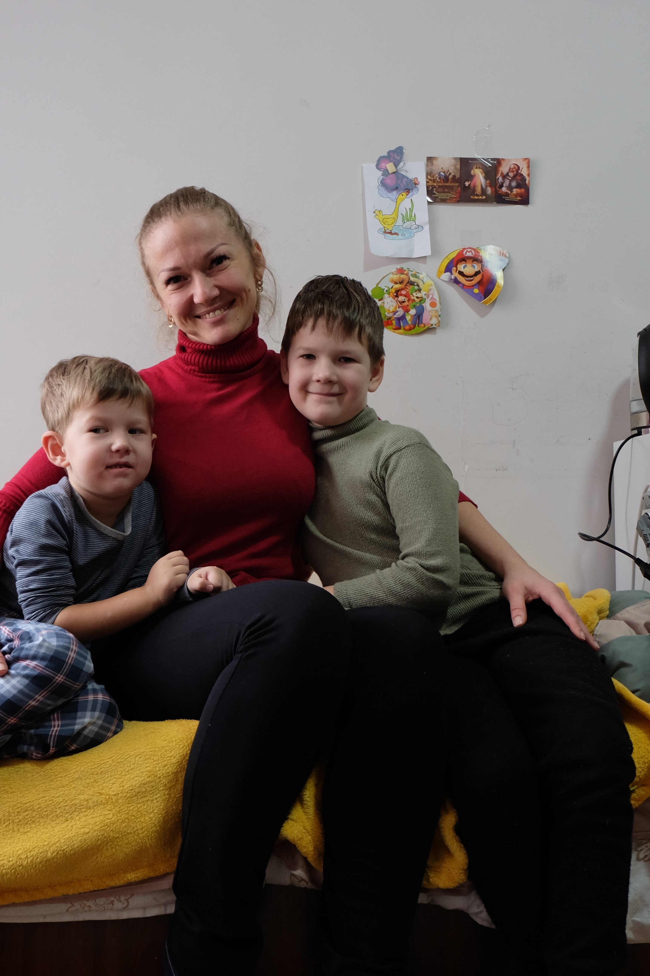 En mamma med sina två barn sitter på en säng. De tittar in i kameran och ler.