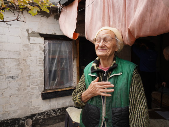 Nadiia, en äldre kvinna i Ukraina, står framför sitt enkla hem.