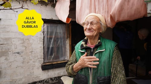 En äldre kvinna står utanför sitt hus. Hon har ena handen på hjärtat och blundar.