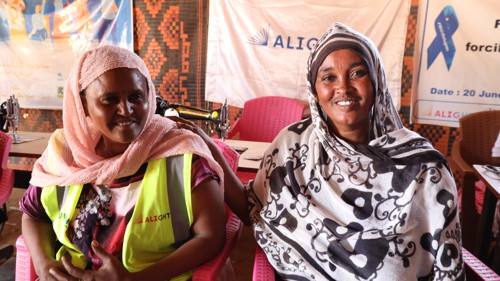 Två etiopiska kvinnor i ett flyktingläger i Sudan sitter på en säng och ler mot kameran.