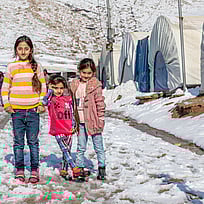 Tre flickor står i ett tältläger i Irak. De har fått varma kläder för att överleva vintern. Du kan hjälpa med ett gåvobevis som ger vinterkläder till barn på flykt.