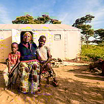 En mamma och två barn sitter framför en nödbostad och tittar in i kameran.