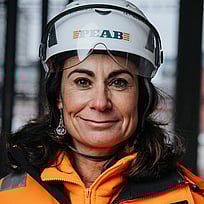 En kvinna klädd i byggkläder med hjälm. Hon tittar in i kameran.