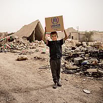 En pojke går längs en grusväg. Han håller en karton över huvudet, på kartongen står det UNHCR.