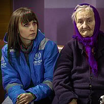 Liudmila, en äldre ukrainsk kvinna, sitter på en säng. Bredvid henne sitter en UNHCR-medarbetare i blå väst.