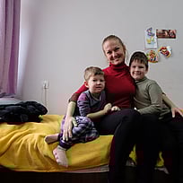 En kvinna och hennes två barn sitter på en säng och ler mot kameran.