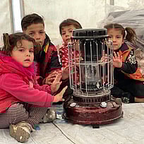 Barn på flykt sitter framför en värmekamin i sitt tält. Du kan hjälpa med ett gåvobevis som värmer familjer på flykt.