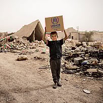 En pojke bär ett nödpaket på huvudet på en tom gata i Syrien. Köp gåvobevis Nödpaket och rädda liv.