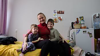 En kvinna och hennes två barn sitter på en säng och ler mot kameran.