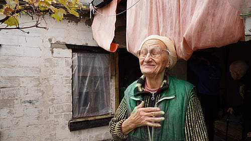 En äldre kvinna står utanför sitt hus. Hon har ena handen på hjärtat och blundar.