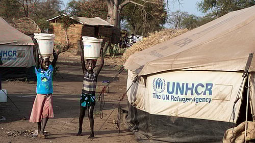 Två barn har vattenhinkar på huvudet och står framför ett tält.