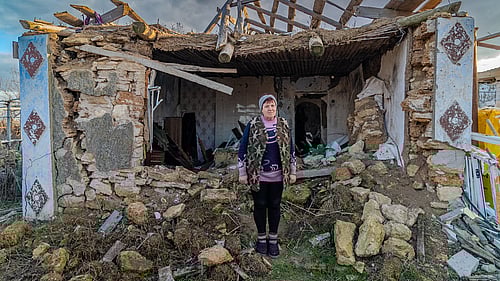 En kvinna står mitt i rasmassorna av sitt sönderbombade hus.