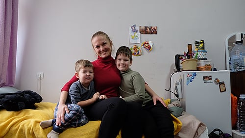 En kvinna med hennes två barn sitter på en säng och ler mot kameran.