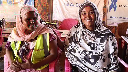 Två etiopiska kvinnor i ett flyktingläger i Sudan sitter på en säng och ler mot kameran.