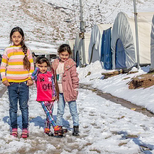 Tre flickor står i ett tältläger i Irak. De har fått varma kläder för att överleva vintern. Du kan hjälpa med ett gåvobevis som ger vinterkläder till barn på flykt.