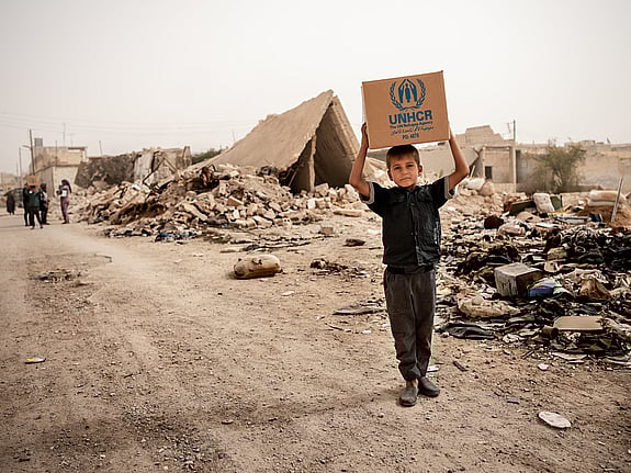 En pojke går längs en grusväg. Han håller en karton över huvudet, på kartongen står det UNHCR.