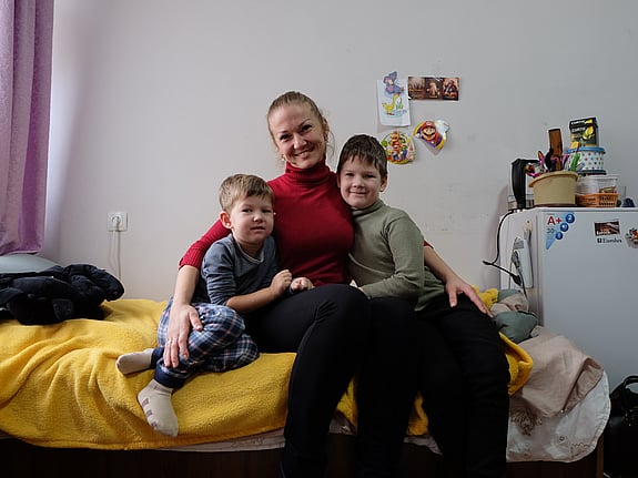 En mamma med sina två barn sitter på en säng. De tittar in i kameran och ler.