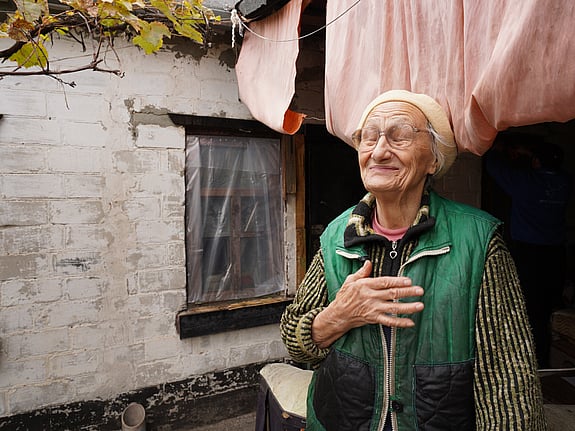 Nadiia, en äldre kvinna i Ukraina, står framför sitt enkla hem.