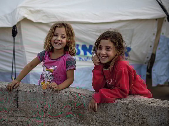 Två flickor i Irak. De ler mot kameran och står framför ett tält.