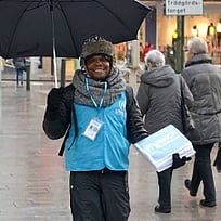 En kille står med ett paraply och UNHCR-väst på ett torg med människor.