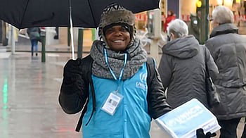 En kille står med ett paraply och UNHCR-väst på ett torg med människor.