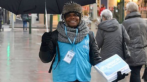 En kille står med ett paraply och UNHCR-väst på ett torg med människor.