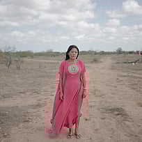 En kvinna i rosa klänning står vid en ödslig väg. Hon tittar in i kameran och ser allvarlig ut.