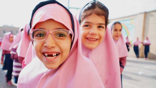 Skolflickor ler framför kampen i en skola i Iran. De två tonårssystrarna flydde från Afganistan till Iran för tio år sedan.   När talibanerna hotade att röva bort flickor som gick i skolan bestämde sig deras pappa Besmellah att det var dags att fly.  2015 tillät regeringen i Iran alla barn att gå i skolan. För första gången fick flickorna en formell utbildning.