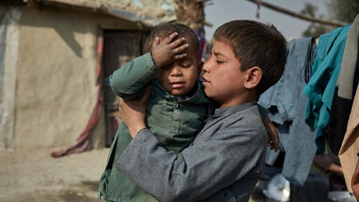 En pojke håller sin lillebror i famnen utanför deras hem. Familjen är på flykt i Afghanistan.